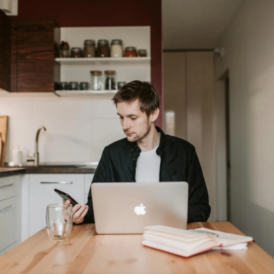 Man sitting at a kitchen table looking at his phone, a laptop is on the table in front of him